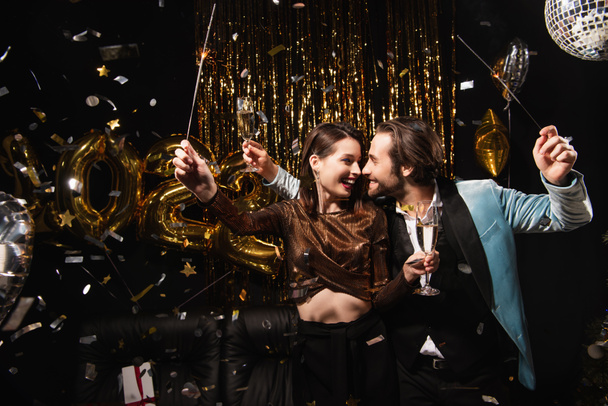 szczęśliwa para z iskrami i szampanem uśmiechająca się do siebie podczas noworocznej imprezy na czarno - Zdjęcie, obraz
