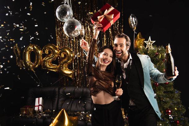 excited woman holding christmas present during festive party with elegant man on black - Photo, Image
