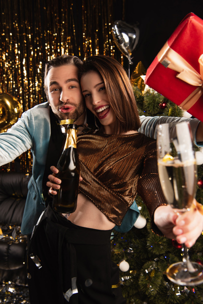 femme heureuse tenant verre et bouteille de champagne près de l'homme ravi avec cadeau de Noël sur noir - Photo, image