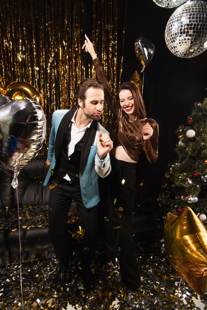 couple excité et élégant dansant près de décoration de Noël brillant sur noir - Photo, image