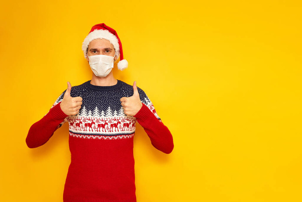 man in Christmas sweater with reindeer, medical mask, New Years hat, a Santa Claus hat, gives a thumbs up, likes, OK sign. isolated yellow background with space for text. holiday concept, gifts, sale - Foto, afbeelding