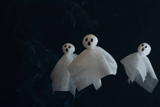 Tres fantasmas blancos hechos de servilletas textiles en el fondo negro con smog blanco, fotografía de stock para cartel de Halloween con espacio para copias. - Foto, Imagen