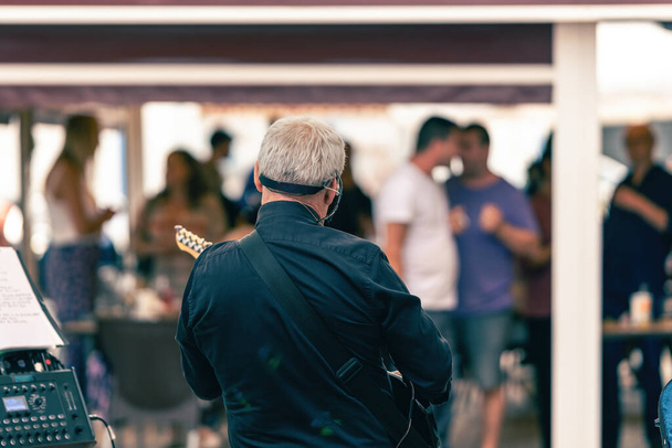 Achterzijde van een oude man die de elektrische gitaar bespeelt op een terras met publiek - Foto, afbeelding