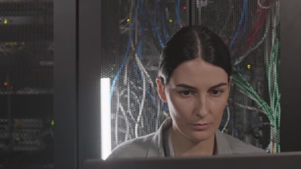 Shoulder-up of brown-eyed Caucasian female engineer leaning back on racks in server room, working, then looking up on camera - Footage, Video