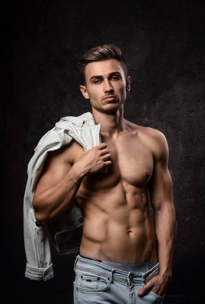 European athletic young man, posing without outerwear, in sports physical form. On a dark textured background. Jacket on the shoulder. - Fotografie, Obrázek