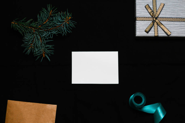 Defocus blank white card with spruce tree branch, gift bow, craft envelope and blue ribbon on black background. Frame mockup, mock up of empty sheet of paper. Xmas holiday composition. Out of focus. - Photo, image