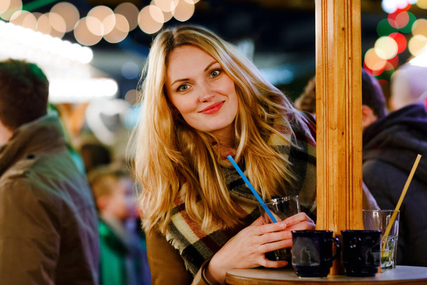 Счастливая молодая женщина пьет горячий пунш, глинтвейн на немецком рождественском рынке. Счастливый человек в зимней одежде с огнями и друзьями на заднем плане. Семья, традиции, концепция праздника. - Фото, изображение