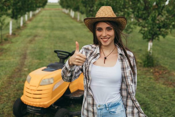 woman posing beside lawn mower in orchard, holding thumb up - Foto, imagen