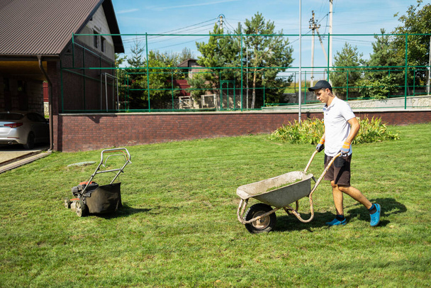 Homem está dirigindo um carrinho de mão em que ele vai coletar grama cortada de um cortador de grama - Foto, Imagem