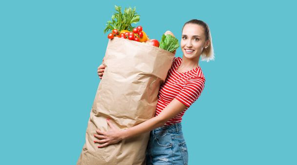 Cheerful woman holding a huge grocery shopping bag filled with fresh vegetables, she is smiling at camera - Fotografie, Obrázek