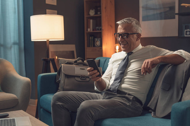 Corporate businessman relaxing on the sofa at home, he is smiling and connecting online using his smartphone - Foto, Imagem