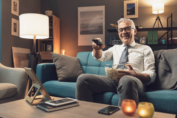 Happy businessman relaxing on the sofa at home: he is watching tv, holding the remote control and eating popcorn - Φωτογραφία, εικόνα