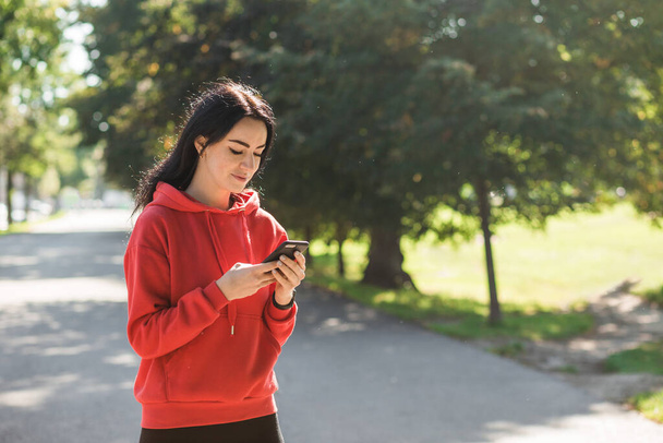 Stílusos lány piros pulóverben pózol egy sikátorban okostelefonnal. Boldog nő gyönyörű mosollyal csevegés mobiltelefon, miközben pihentető a parkban, szabadban - Fotó, kép