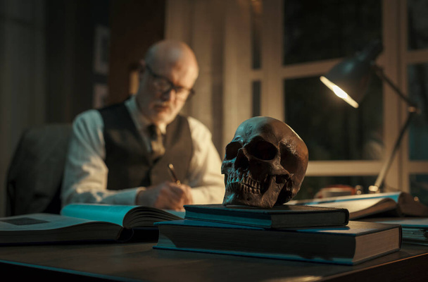 Työpöydän ääressä työskentelevä professori ja ihmisen kallo etualalla, tietämyksen ja tutkimuksen konseptissa - Valokuva, kuva