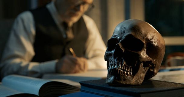 Academic professor sitting at office desk, he is studying a human skull and writing a report - Фото, зображення