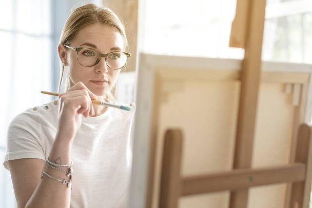 Young woman with glasses painting on canvas in the studio - Photo, Image