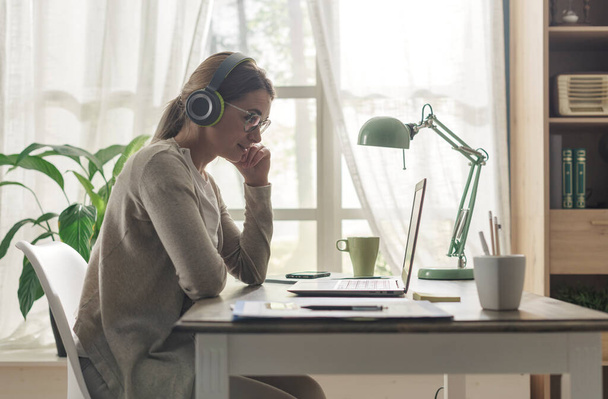 Young woman sitting at desk and connecting with her laptop, she is wearing headphones and watching videos online - 写真・画像
