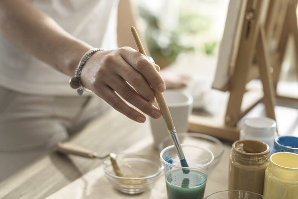 Creative woman painting in the atelier, she is dipping the paintbrush in a glass - Foto, Bild