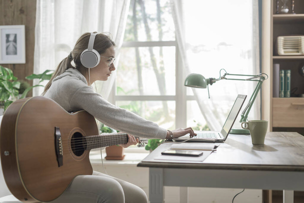Mujer joven sentada en el escritorio en casa, está tocando la guitarra y conectándose en línea con su computadora portátil - Foto, imagen