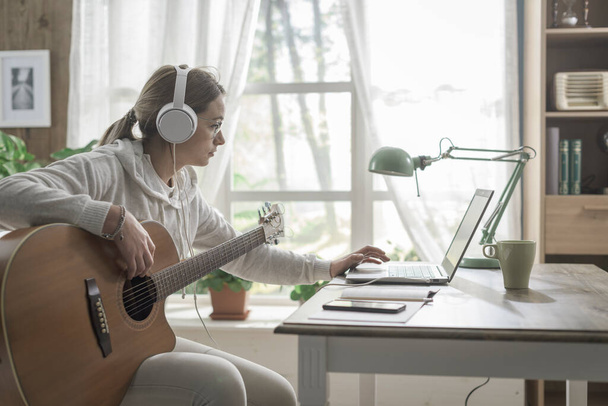 Mujer joven sentada en el escritorio en casa, está tocando la guitarra y conectándose en línea con su computadora portátil - Foto, imagen