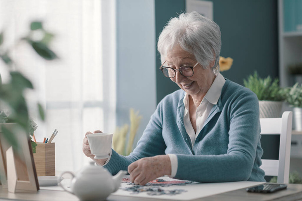 Donna anziana felice rilassarsi a casa da sola, sta risolvendo un puzzle e bere il tè - Foto, immagini