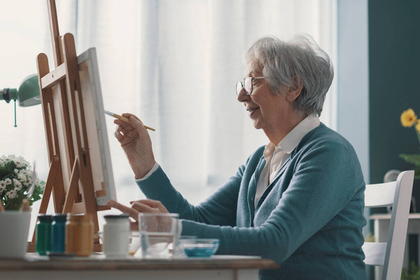 Happy senior woman sitting at desk at home and painting on canvas - Fotó, kép