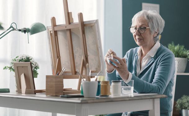 Happy senior woman sitting at desk at home and painting on canvas - Фото, зображення