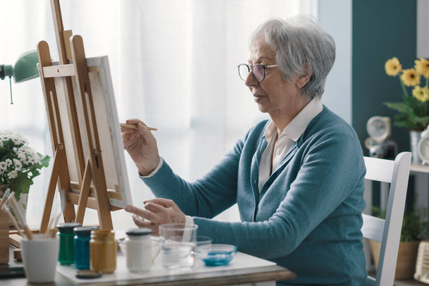 Happy senior woman sitting at desk at home and painting on canvas - Photo, Image