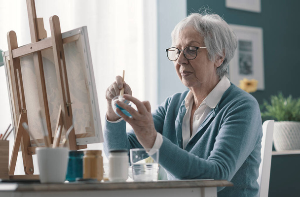 Happy senior woman sitting at desk at home and painting on canvas - Foto, afbeelding