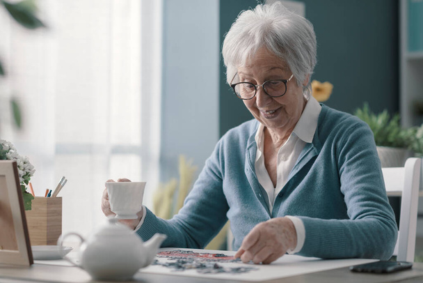 Happy elderly woman relaxing at home alone, she is playing puzzle and drinking tea - Foto, imagen