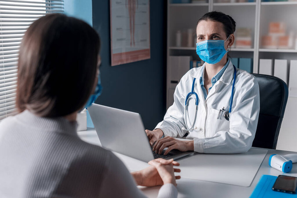 Female doctor meeting a patient in her office for a medical consultation, they are wearing surgical masks - Фото, зображення