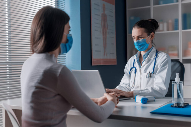 Female doctor meeting a patient in her office for a medical consultation, they are wearing surgical masks - Valokuva, kuva