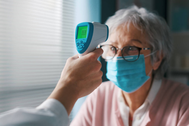 Médico verificando a temperatura corporal de um paciente sênior usando um termômetro infravermelho sem contato - Foto, Imagem