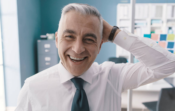 Friendly corporate businessman standing in his office, he is laughing and touching his head - Foto, Imagem