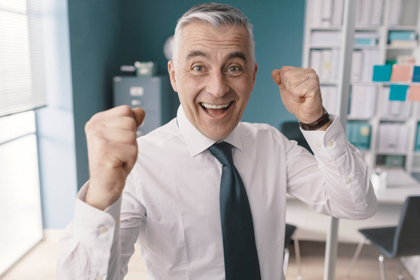 Cheerful smiling businessman celebrating his success in the office with raised fists - Photo, image