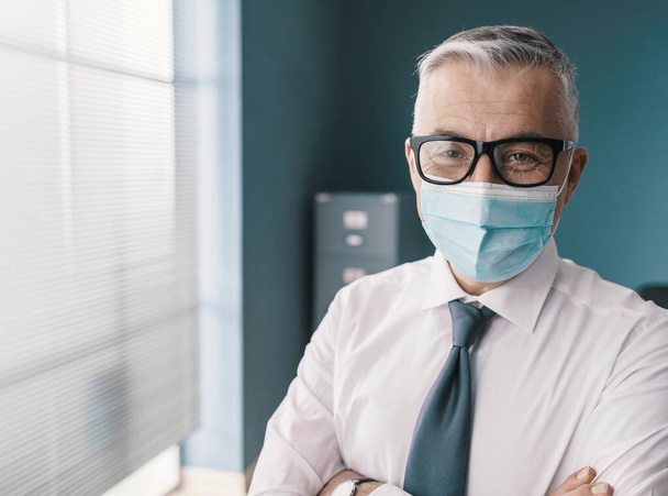Confident corporate businessman posing in the office, he is wearing a surgical mask and glasses - 写真・画像