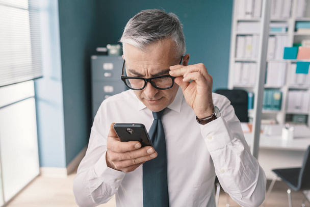 Corporate businessman using his smartphone and having vision problems, he is adjusting his glasses - Foto, Imagem