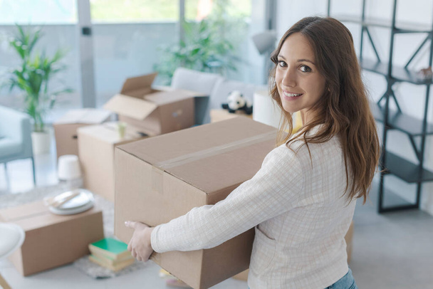 Happy young woman moving in her new home, she is carrying a cardboard box with her belongings - Фото, изображение