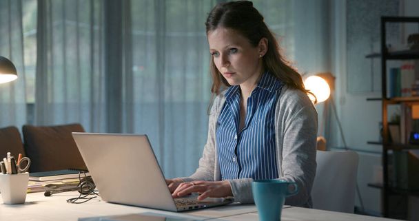 Young woman sitting at desk and working from home, she is typing on her laptop - Fotó, kép