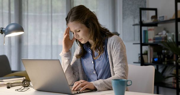 Young woman sitting at desk at home and feeling tired and stressed, she is having a bad headache - Фото, изображение