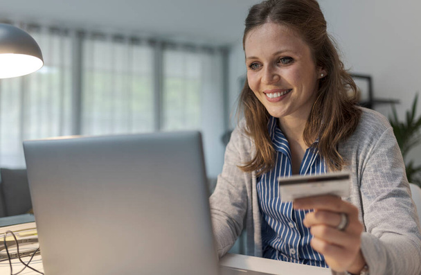 Χαμογελώντας νεαρή γυναίκα κάθεται στο γραφείο και χρησιμοποιώντας μια πιστωτική κάρτα σε απευθείας σύνδεση, κάνει online ψώνια - Φωτογραφία, εικόνα
