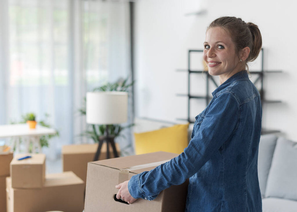 Happy young woman carrying a box in her new apartment and smiling, home relocation concept - Fotó, kép