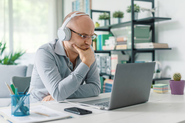 Empresario sentado en el escritorio y trabajando desde casa, lleva auriculares y se conecta con su computadora portátil - Foto, Imagen
