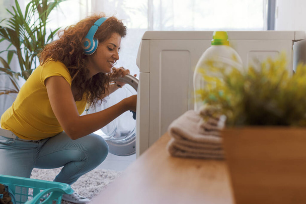 Happy young woman doing laundry at home, she is loading the washing machine and listening to music - Foto, afbeelding