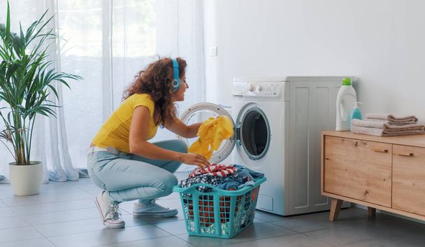 Happy young woman doing laundry at home, she is loading the washing machine and listening to music - Φωτογραφία, εικόνα
