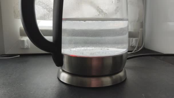 Kookwater in een transparante theepot op de keukentafel bij het raam. Luchtbellen rijzen op in een glas, elektrisch keukenapparaat. Koffiepauze en einde van het werkdagconcept. - Video