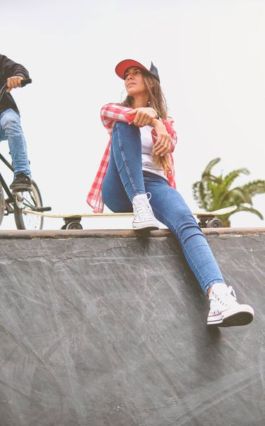 Schöne junge Frau mit Mütze sitzt mit ihrem Longboard im Skatepark auf der Rampe. - Foto, Bild