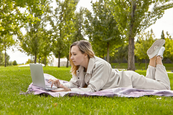 The woman is lying down using a laptop in the park. Smiling young student woman is using a laptop laying on grass. The concept of teaching in the park. Student woman relaxes while sitting on a blanket in park.  - Foto, Bild