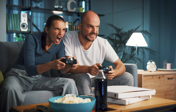 Couple sitting on the couch at home and playing video games together, they are holding the controller and focusing on the game - 写真・画像