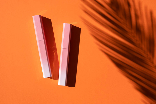 Brillo labial en una caja degradada de color rosa a blanco con sombra de hojas de palma. Vista superior sobre fondo naranja. - Foto, imagen